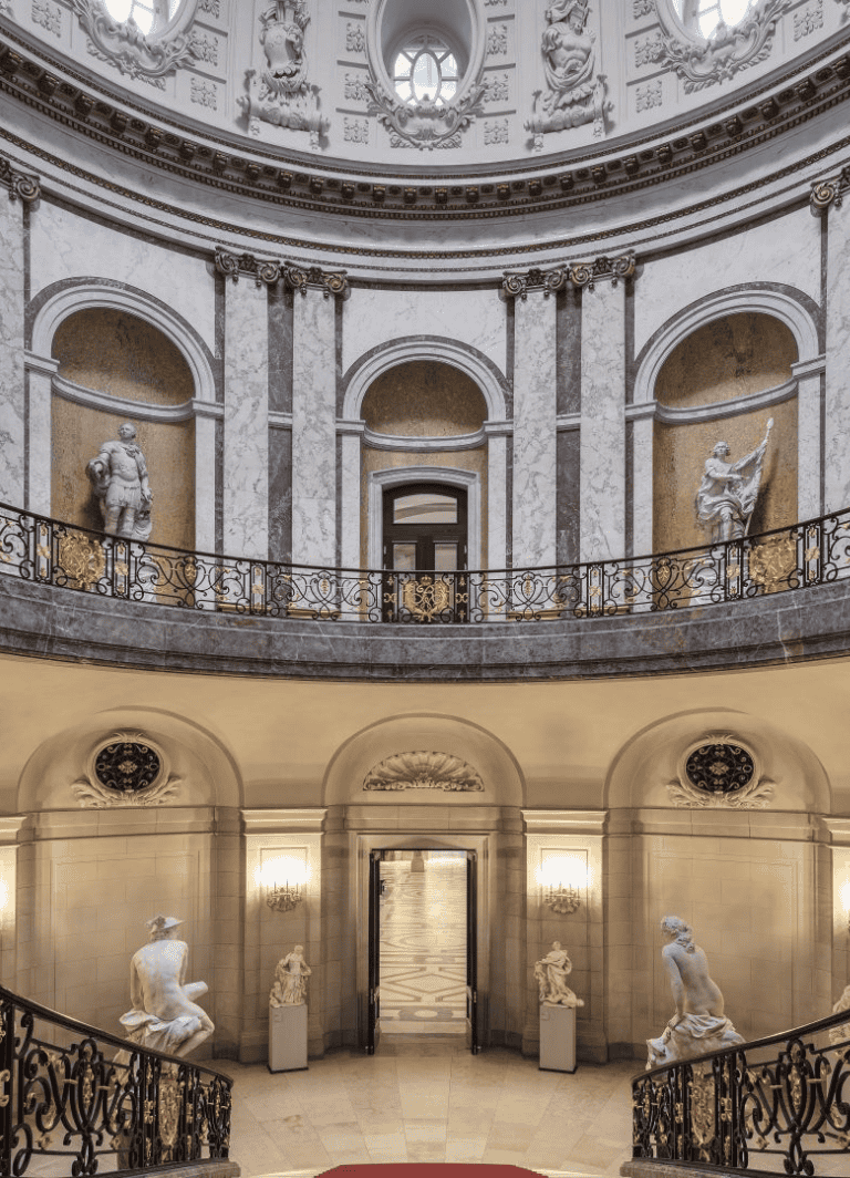 Elegant marble interior with classical statues and ornate iron railings, showcasing grandeur and historical architecture.