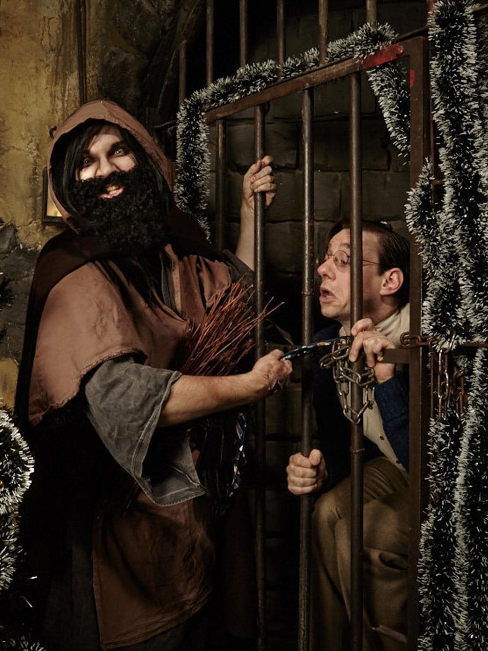 Man dressed as a medieval jailer with humorous prisoner behind bars for holiday themed escape room game.
