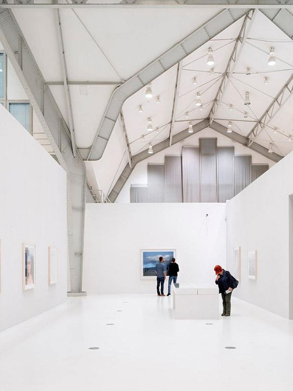 Vibrant art gallery interior with white walls and high ceiling, showcasing paintings and visitors appreciating art.