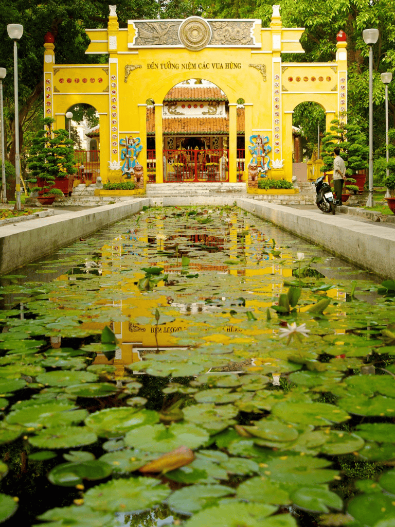 Colorful temple entrance with water lily pond in the foreground, lush greenery, and traditional Vietnamese architecture.