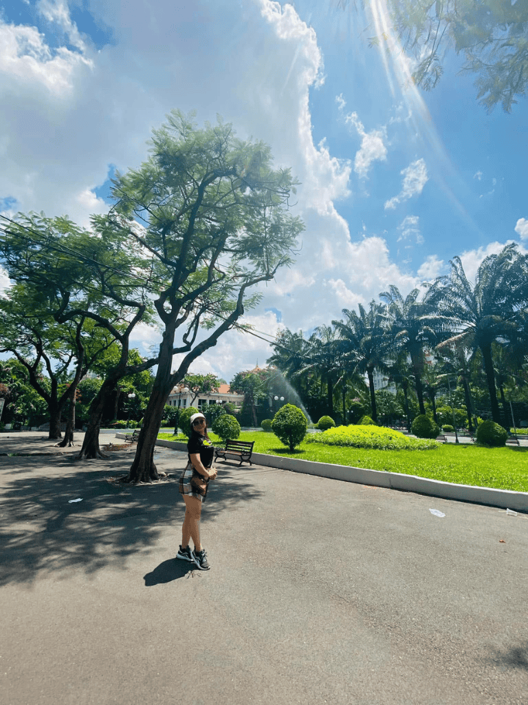 Bright sunny park with lush green trees and a woman in athletic attire enjoying outdoor scenery.