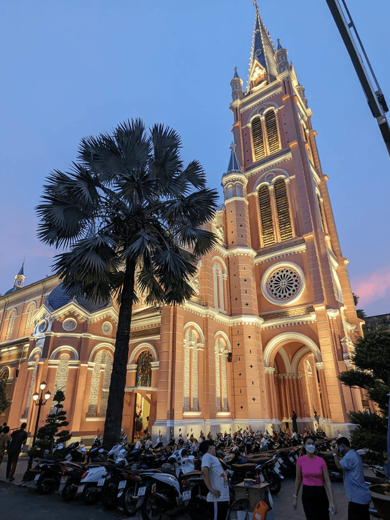 Colorful church illuminated at dusk with palm tree in front, bustling scooter parking and pedestrians nearby.