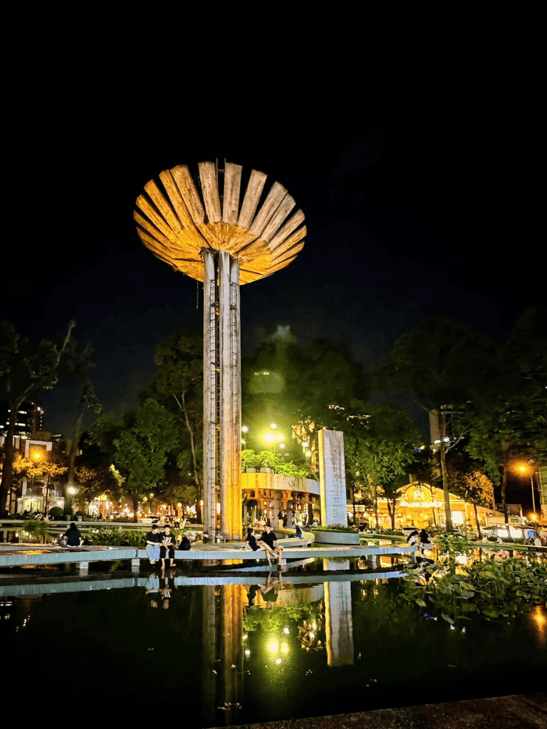 Night view of Quest for Directions landmark with illuminated tower and reflection in water, lively urban park scene.
