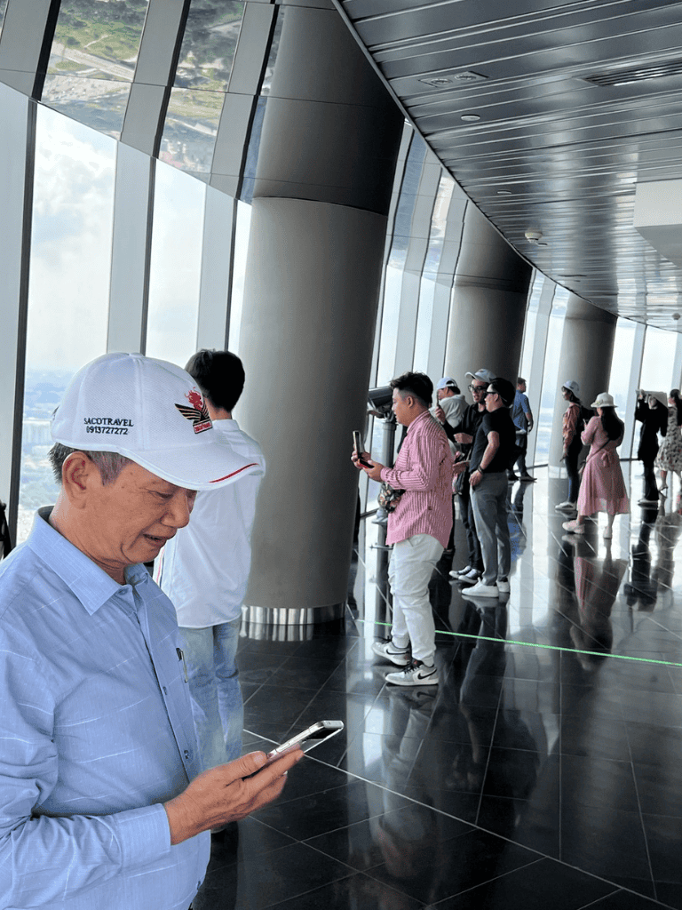 Elevator observation deck with tourists viewing cityscape and using mobile phones.