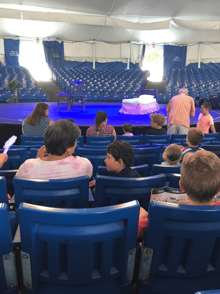 Open-air theater stage with audience waiting, blue seating, and vibrant atmosphere.