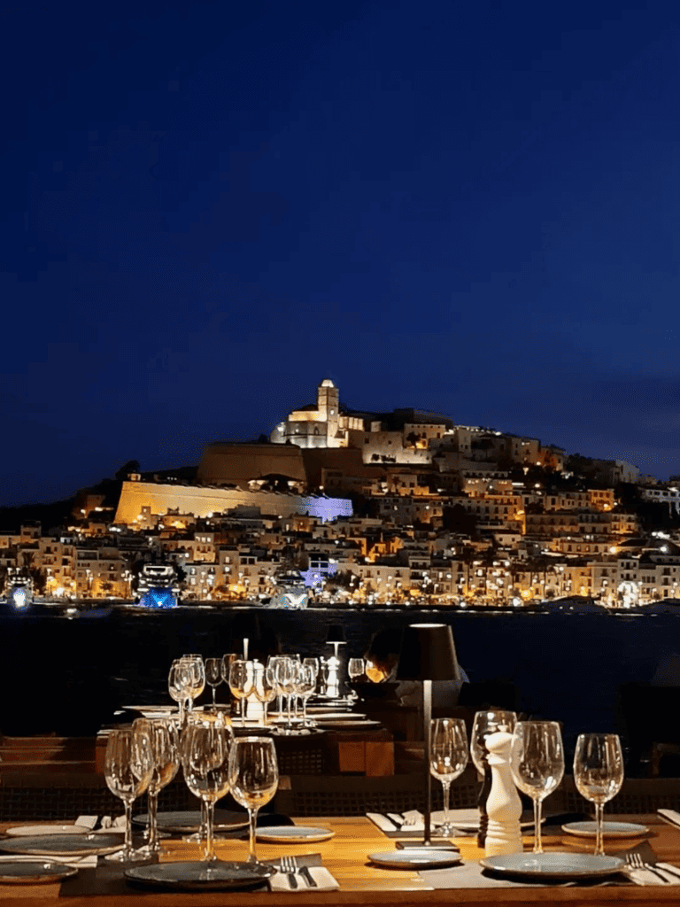 Night view of a coastal city with illuminated buildings and a historic fortress, perfect for travel and dining experiences.
