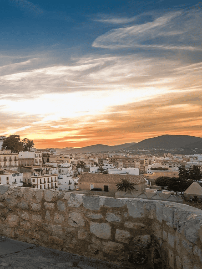 Ancient Mediterranean town at sunset with white buildings and mountain backdrop.