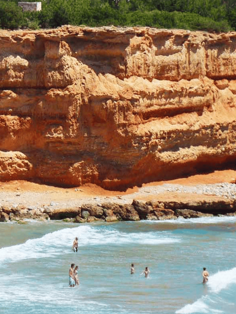 Cliffs by the beach with people swimming and relaxing in clear waters.