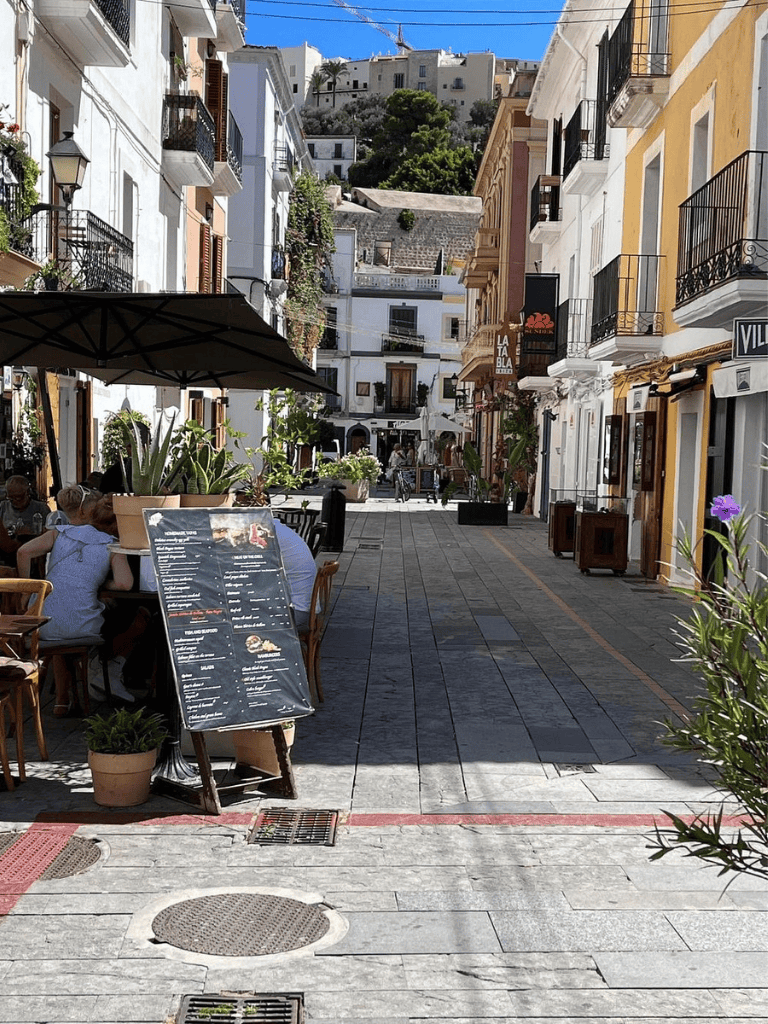 Alto text: Vibrant Mediterranean street with cafes, colorful buildings, and hilltop views.