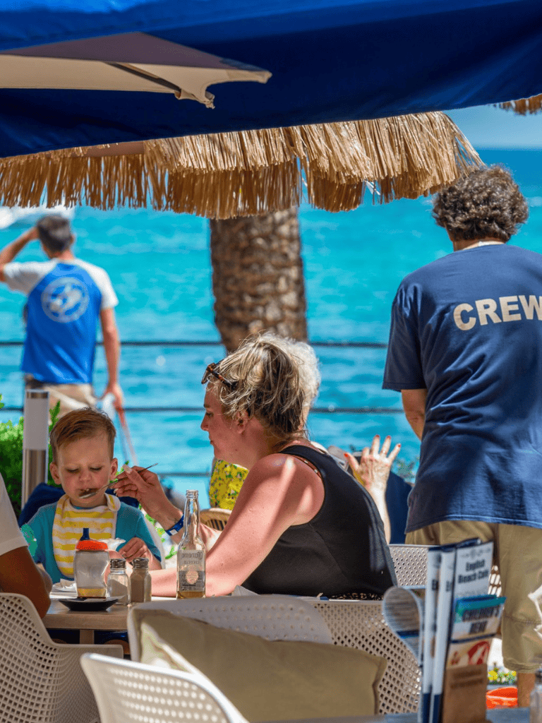 Relaxing beachside scene with family dining and crew member, sunny weather and ocean view.