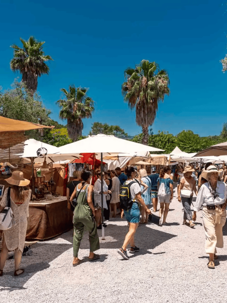 Vibrant outdoor market with palm trees, shoppers, and vendor stalls on a sunny day.