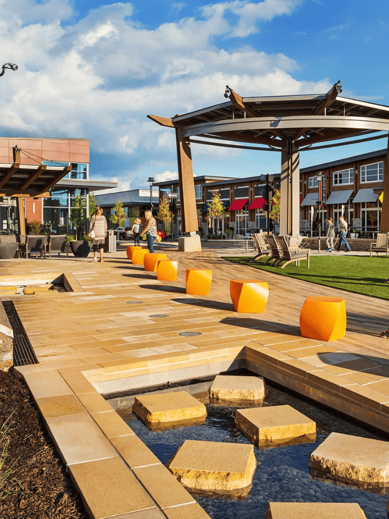 Vibrant outdoor shopping and dining area with modern architecture, seating, and water features in a community plaza.
