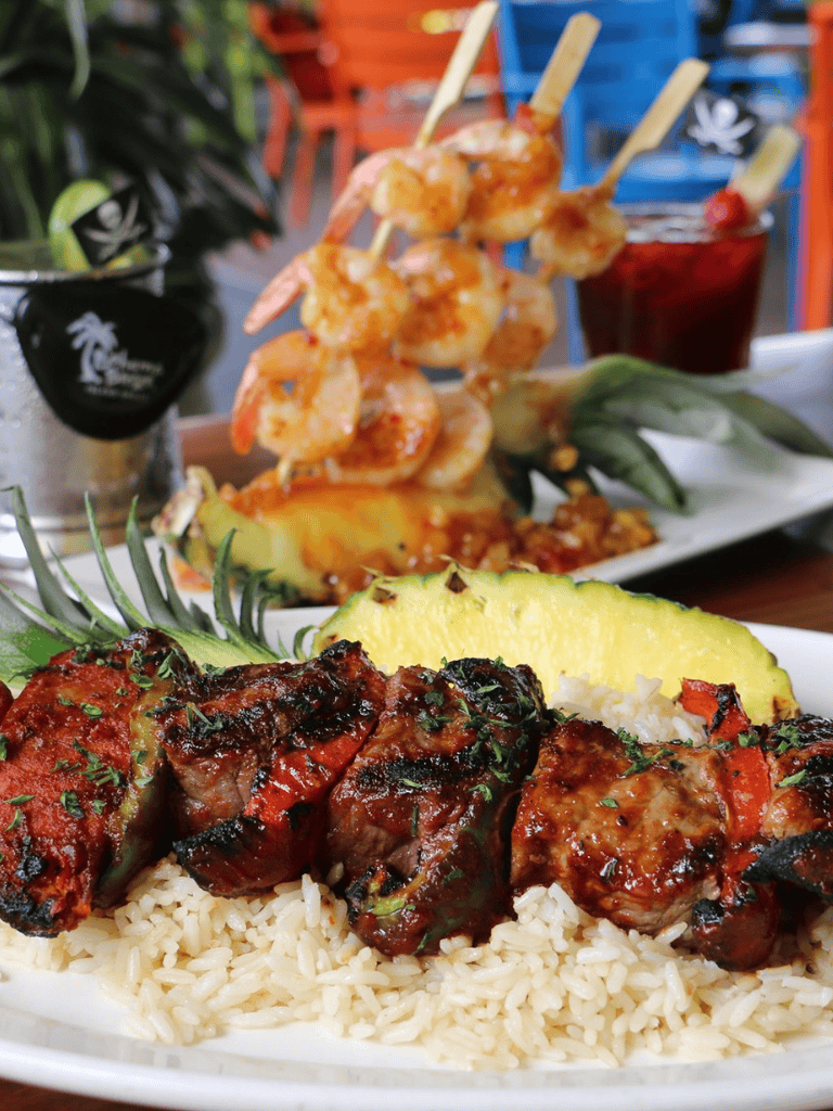 Succulent grilled meat skewers with rice, pineapple, and tropical fruit garnishes at a vibrant restaurant.