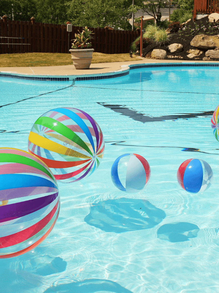 Colorful beach balls floating in a backyard swimming pool with a garden and patio in the background.