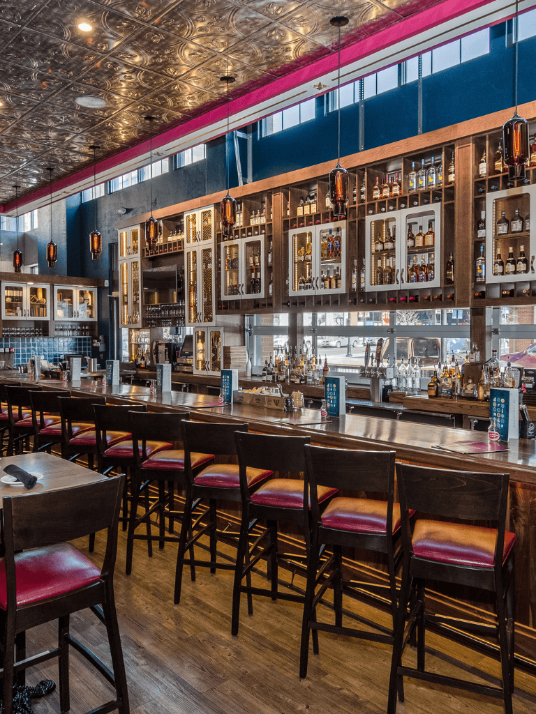 Cozy modern bar interior with liquor bottles, wooden shelves, and high chairs at the counter.