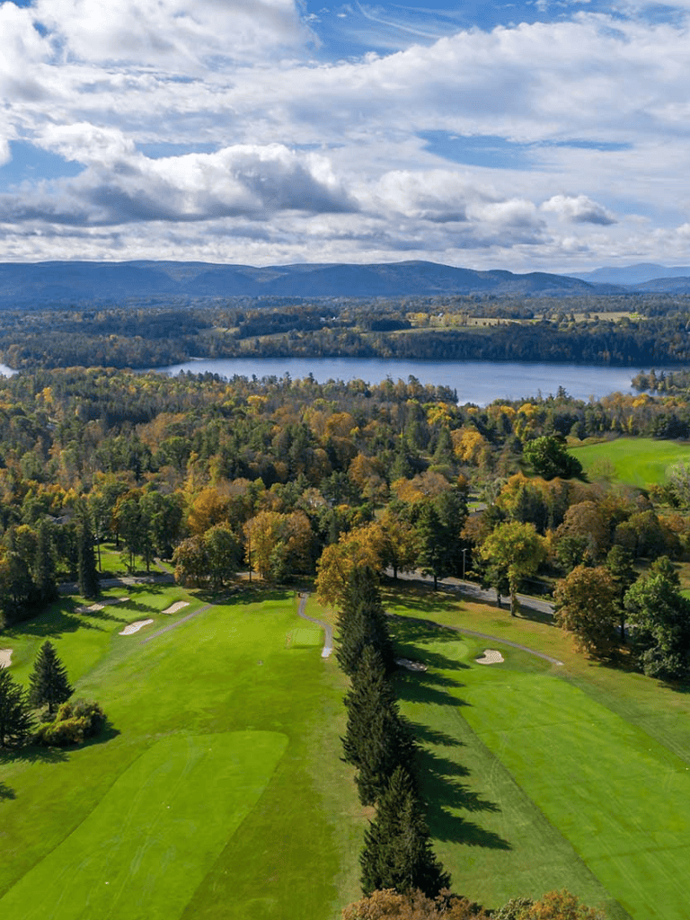 Vibrant golf course surrounded by autumn trees with lake and mountains in background.