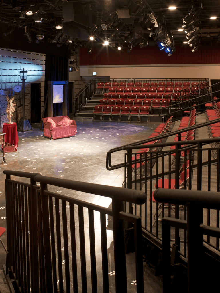 Empty theater-style comedy club with stage, red seats, and private viewing area for performances.