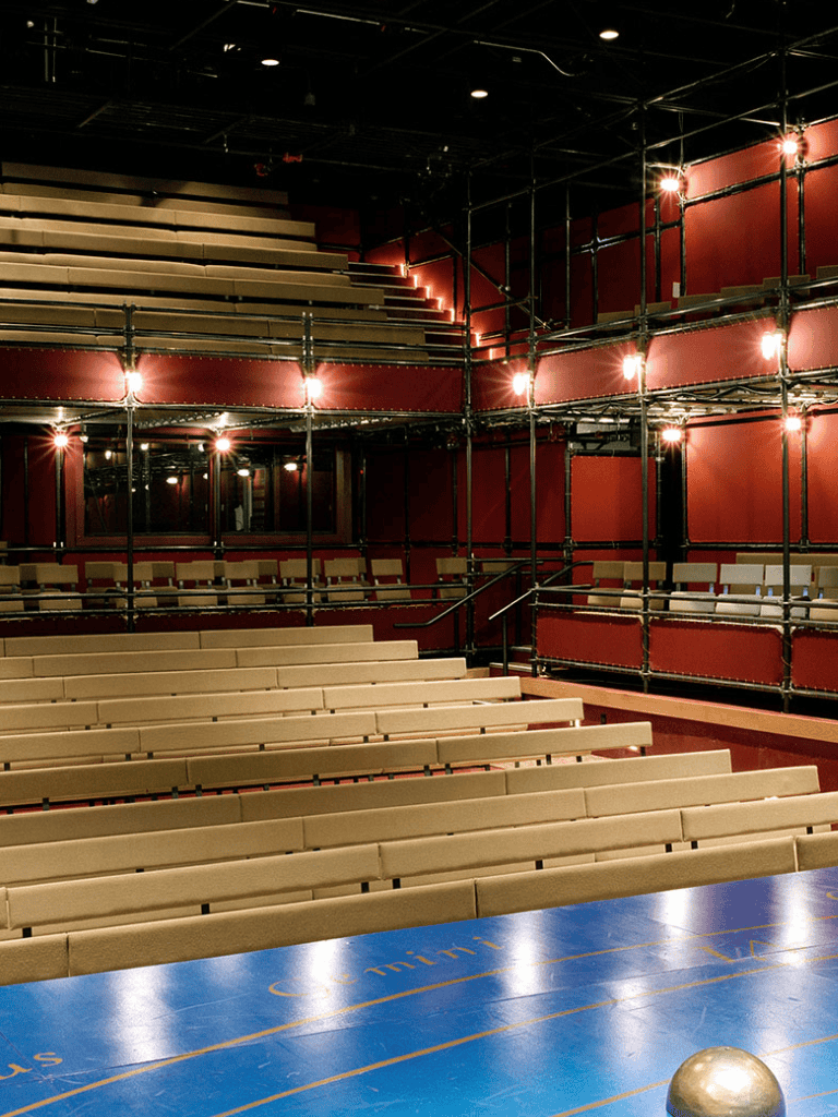 Empty theater with red walls, tiered seating, and stage in the background, illuminated by warm lighting.