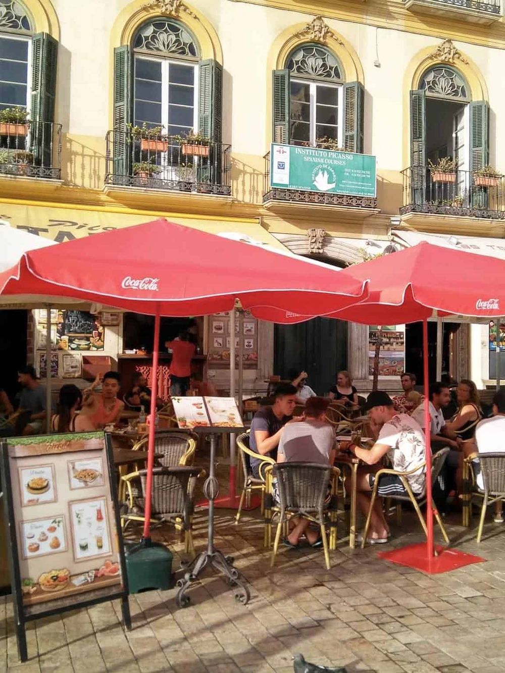 Outdoor restaurant cafe with people enjoying meals under red umbrellas in a vibrant city setting.