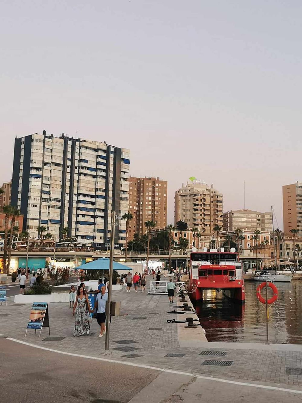 Vibrant city harbor scene featuring tall modern buildings, a red boat docked, and people enjoying waterfront views.