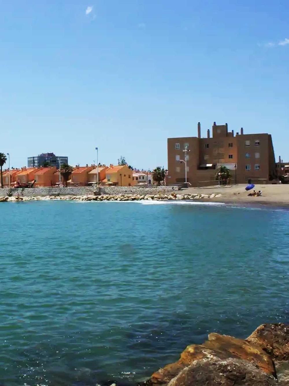 Beachfront residential buildings along a calm ocean shoreline with clear blue sky and rocky coast.