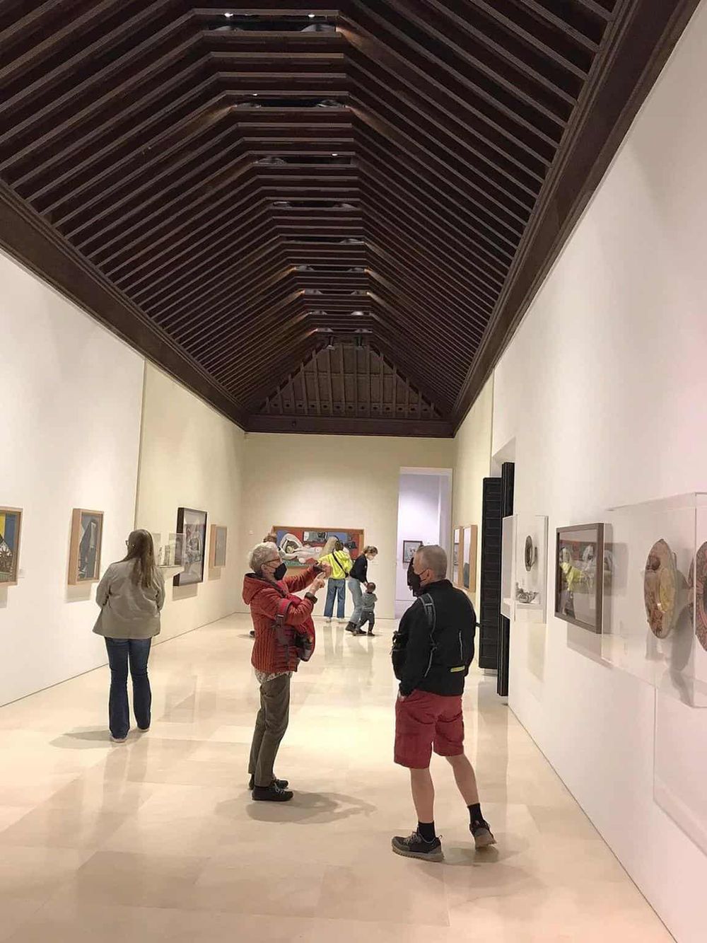 Colorful art gallery interior with visitors viewing paintings and sculptures, modern museum space with high wooden ceiling.