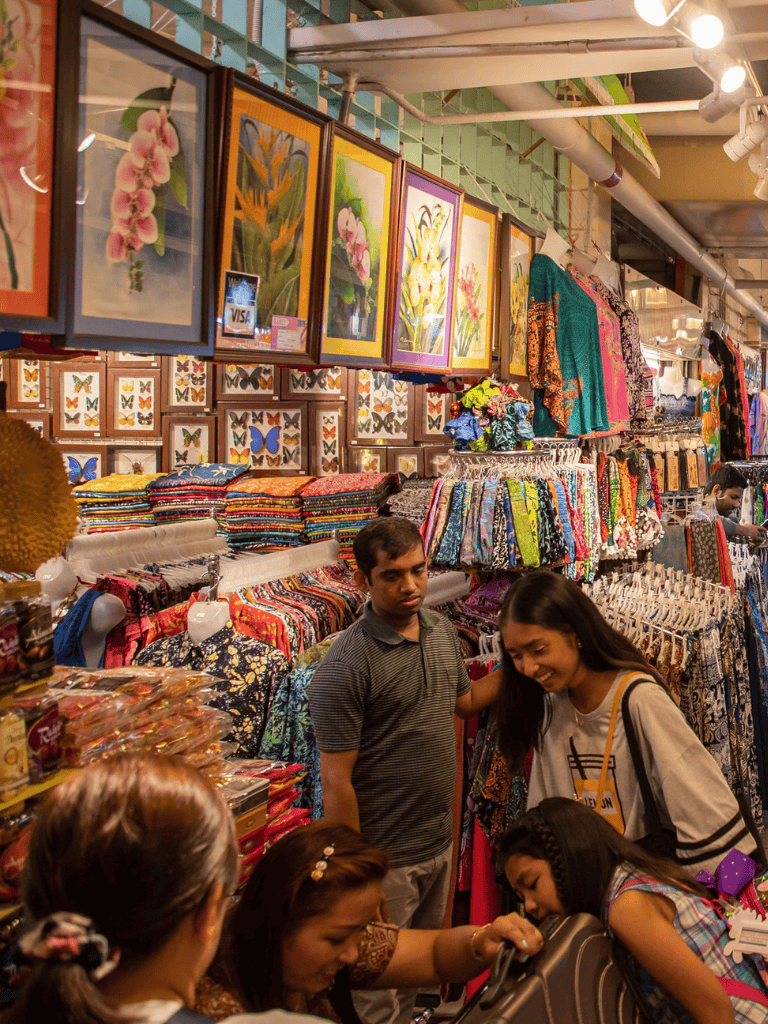 Colorful clothing and art store with shoppers browsing and vibrant displays of fabrics, paintings, and accessories.
