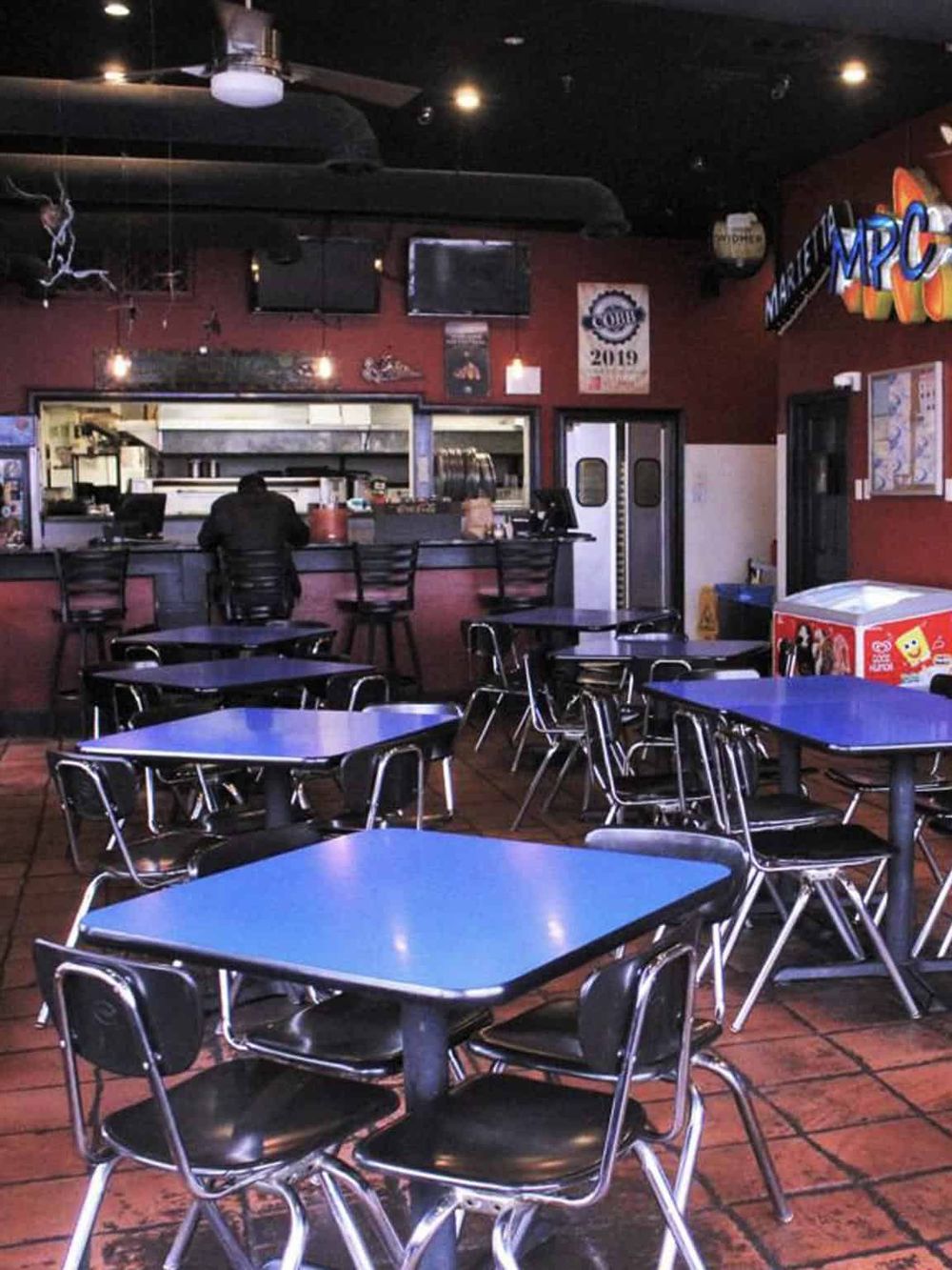 Brightly lit indoor sports bar with multiple tables, TVs, and a visible open kitchen area.