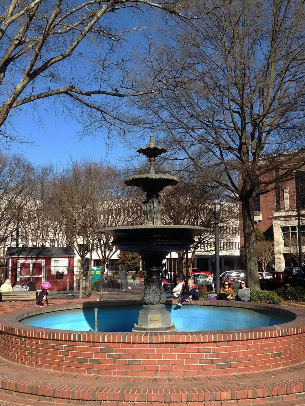 Vintage-style public fountain in a city park with trees and people relaxing on a sunny day.