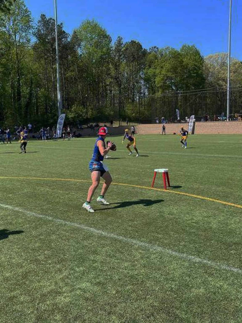 Youth football practice on grassy field, wearing sports gear and participating in drills, sunny day, trees in background.