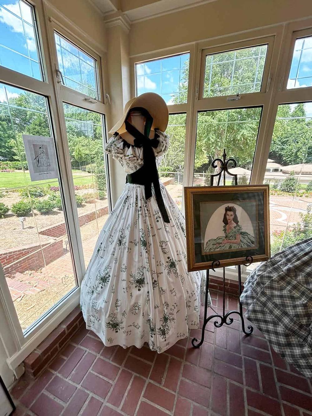 Vintage fashion display featuring a 1950s style dress, hat, and artwork inside a sunlit conservatory.