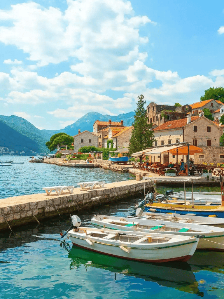 Serene coastal village with boats, historic buildings, mountains, and waterfront dining on a sunny day.