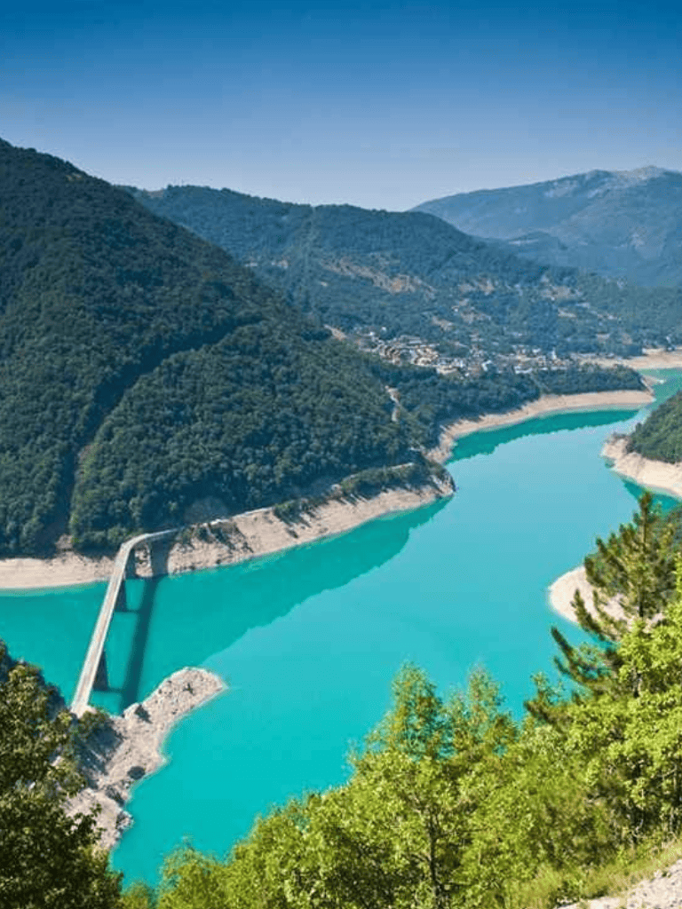 Vivid image of a turquoise dam reservoir nestled in lush green mountains with a bridge crossing the water.