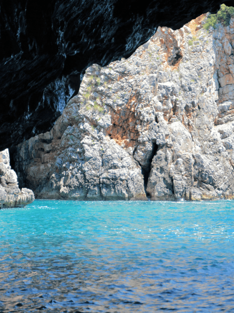 Vibrant turquoise water beneath a rocky, cave-like overhang in a scenic coastal landscape.