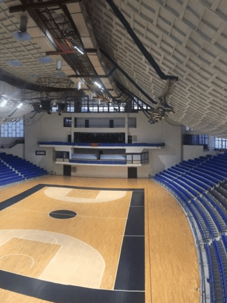Indoor basketball court with seating, scoreboard, and high ceiling.