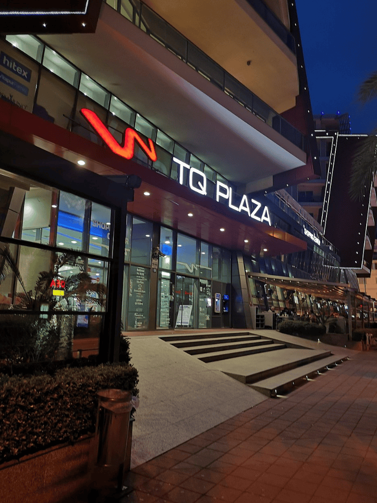 Modern shopping mall exterior at night, illuminated signage for TQ Plaza in a bustling city area.