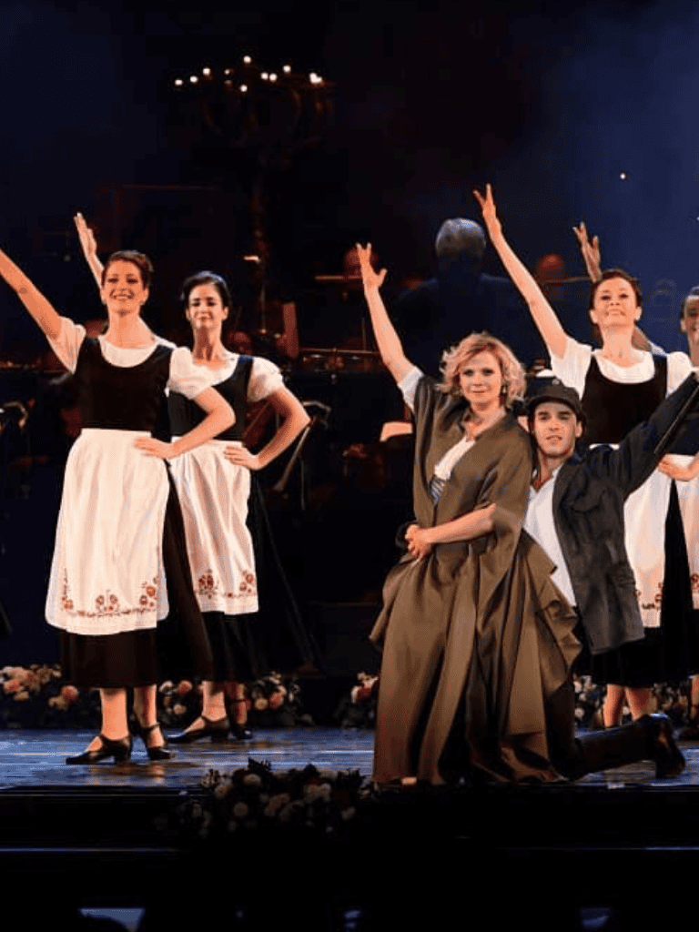 A girl performs traditional folk dance on stage with a group in historical costumes.
