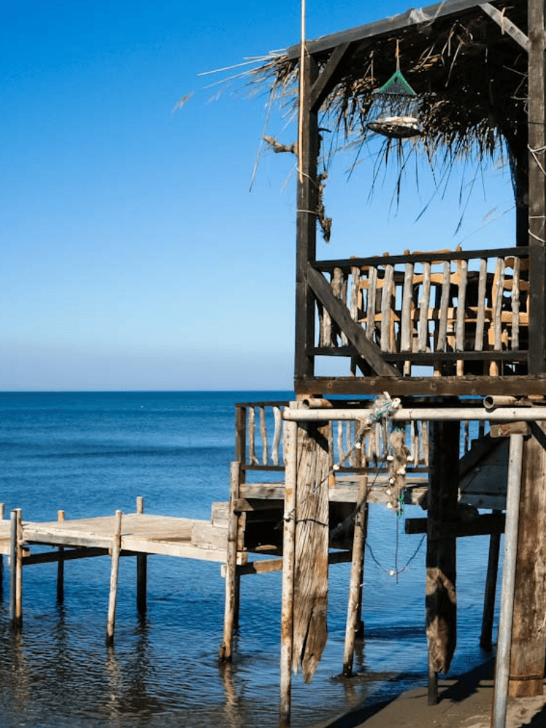 Dilapidated wooden pier over calm ocean waters, tropical hut with thatched roof, sunny day, serene coastal scenery.