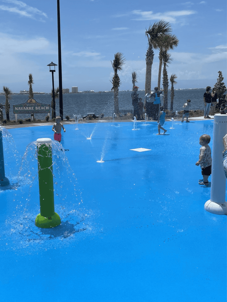 Children enjoying water spray play area at Navarre Beach with ocean view in background.