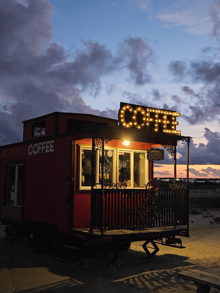 Coffee stand illuminated at dusk on a portable cart with a "CLOSED" sign, on a beach with dramatic clouds.