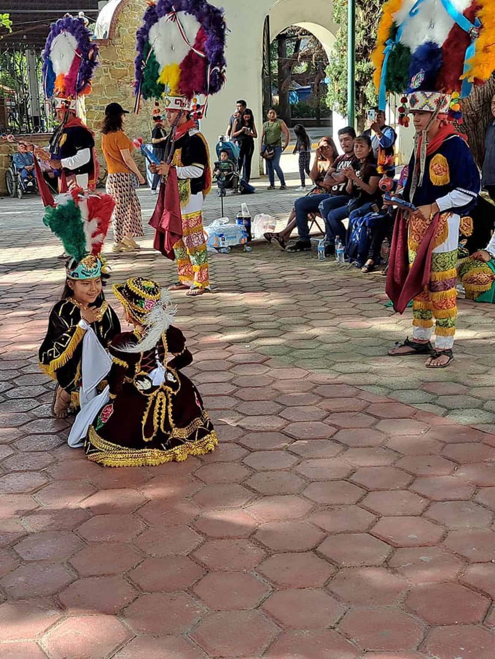 Colorful traditional dancers in cultural costumes, performing at an outdoor event with audience members watching.