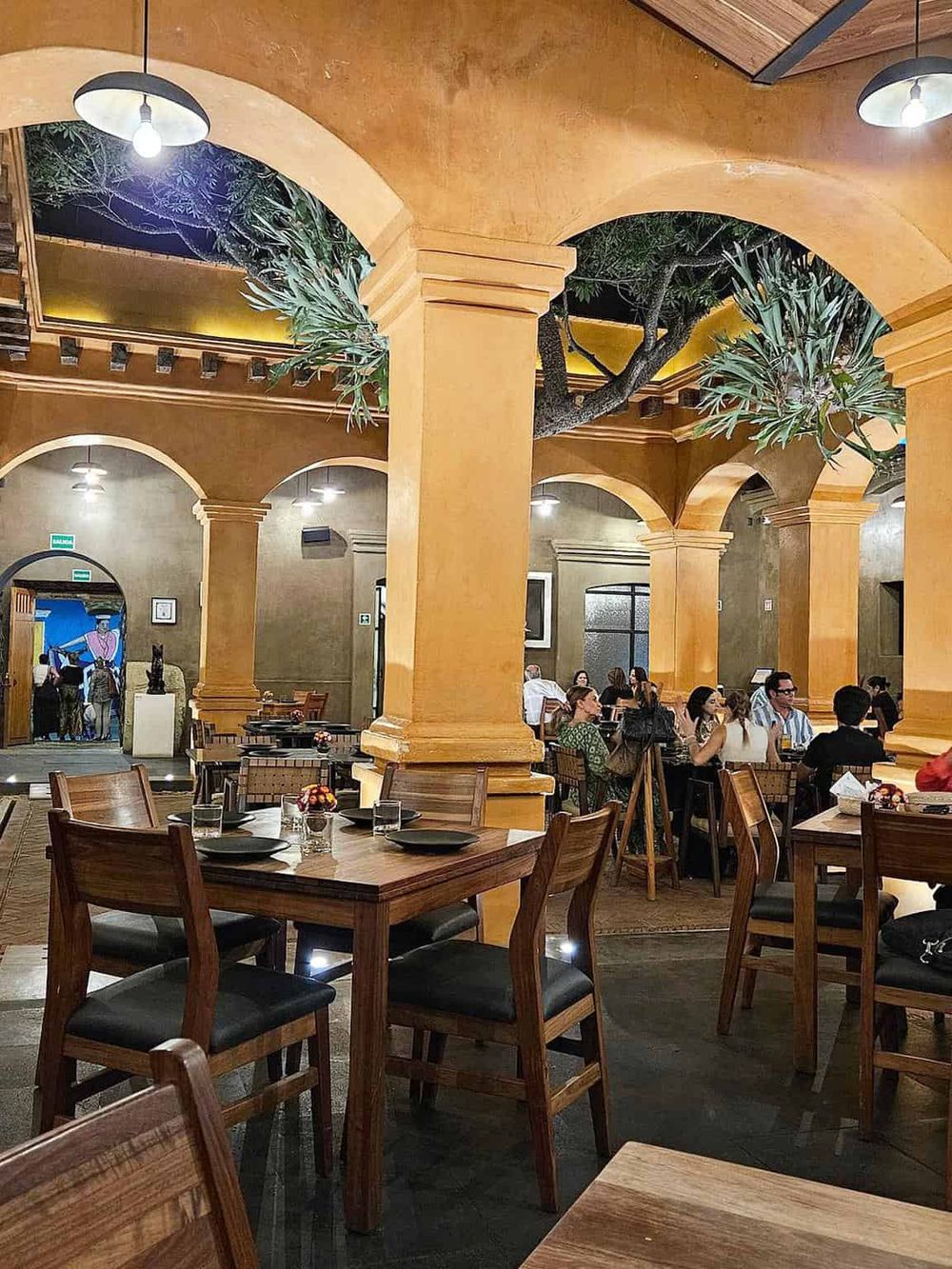 Warm-lit restaurant interior with arches, wooden furniture, and lush greenery, offering a cozy dining experience.