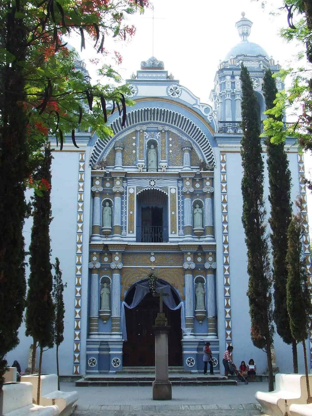 Colorful church with ornate architecture, surrounded by trees and visitors in Mexico City.