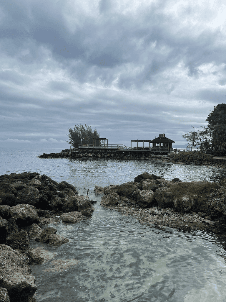 Serene lakeside scene with rocky shore, pavilion, and cloudy sky in a scenic outdoor setting.