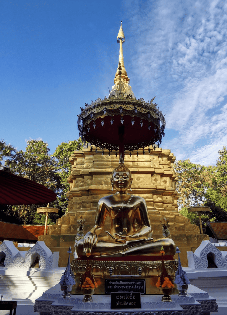 Golden Buddha statue at a Thai temple, with ornate stupa and blue sky background.
