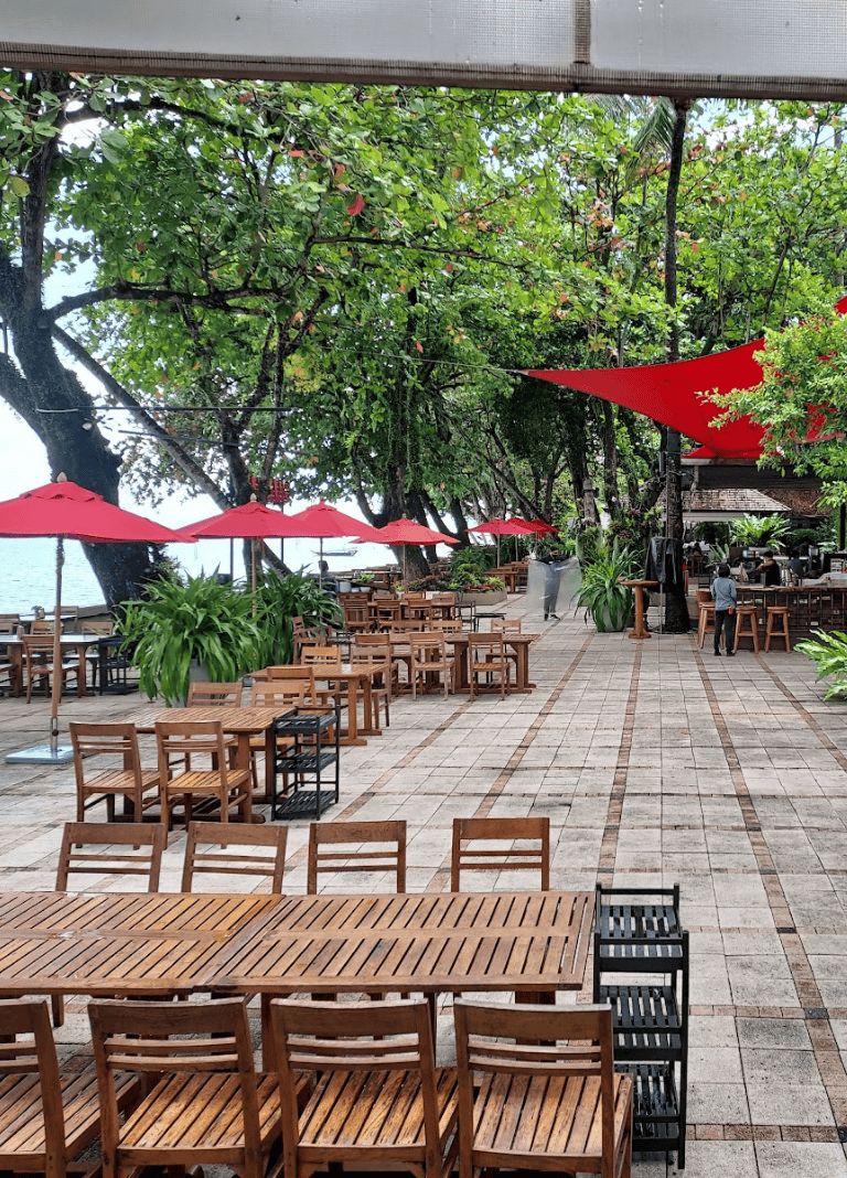 Outdoor waterfront dining area with wooden tables, red umbrellas, lush greenery, and shaded trees.
