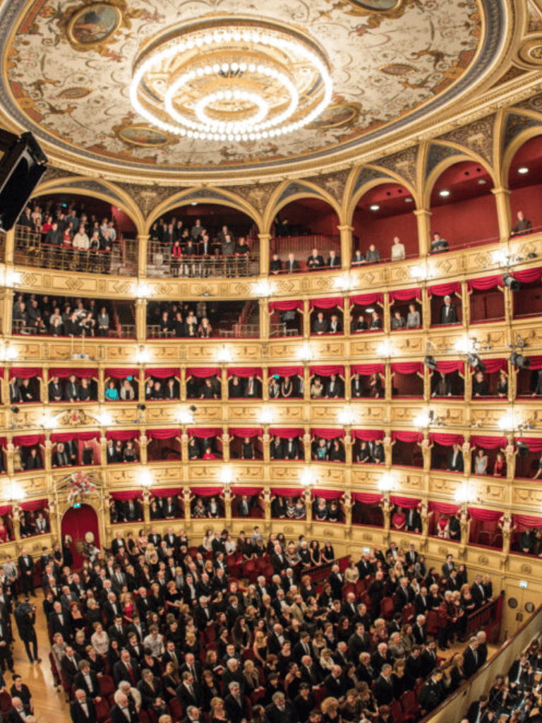 An elegant theater with multiple balconies filled with an audience attending a live performance.