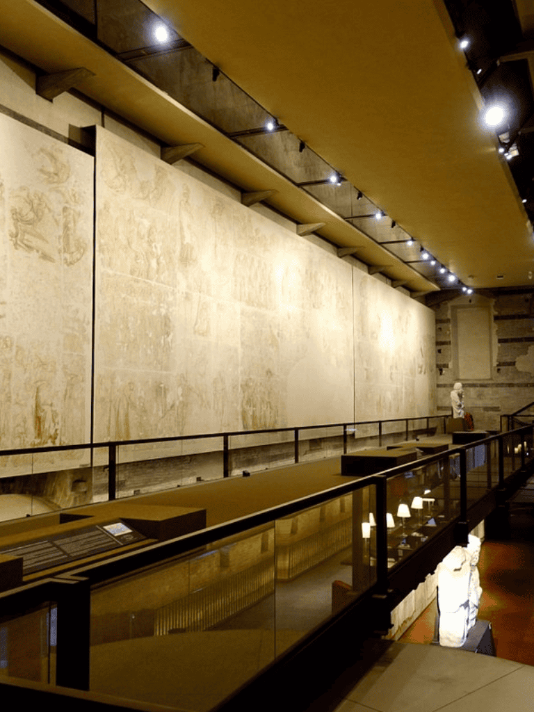 Ancient wall display in a modern museum with lighting and railing, showcasing historical artifacts and artworks.