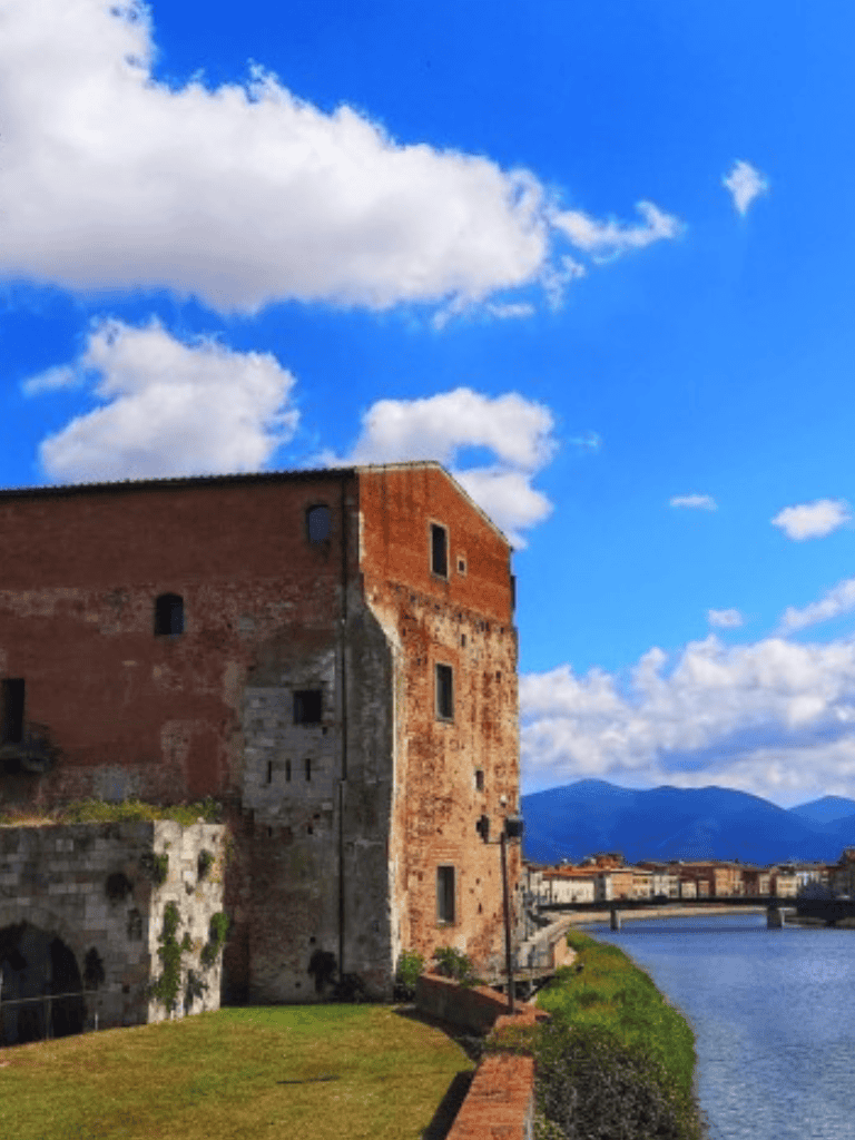 Historic brick building along a riverbank with mountains in the background, showcasing scenic Italian architecture and landscape.