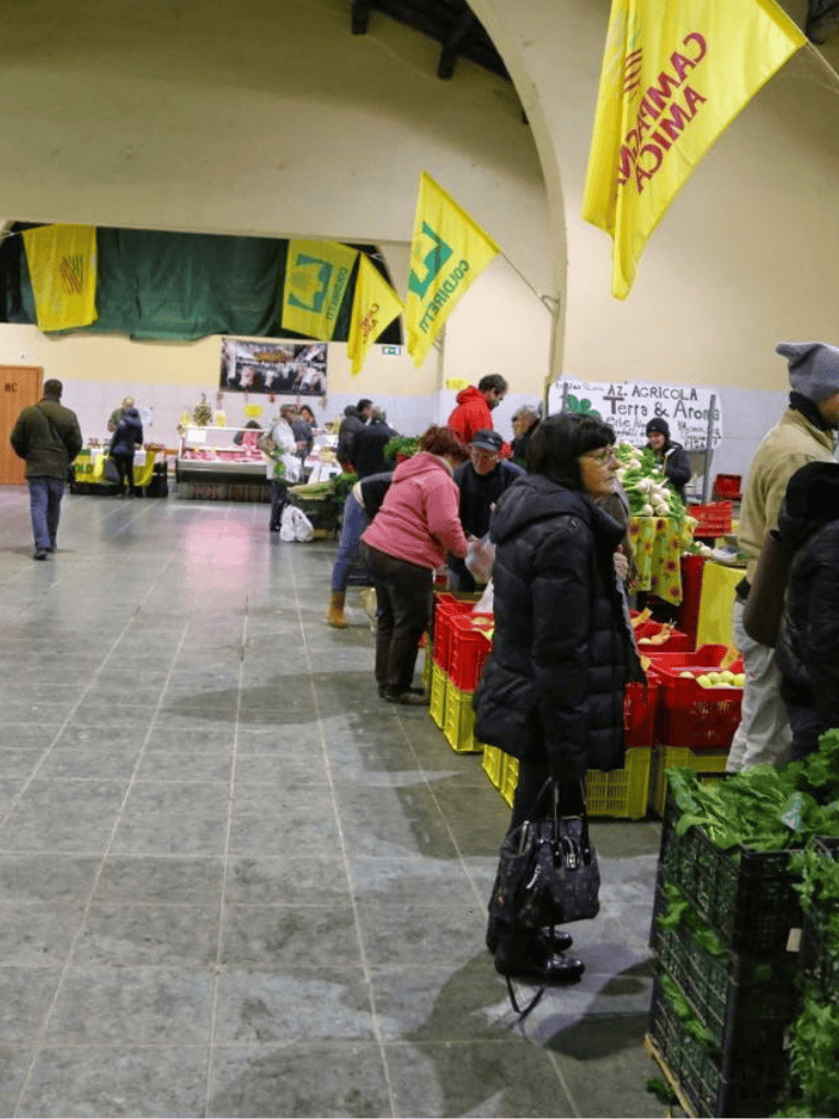Fresh produce at indoor farmers market, visitors shopping for organic fruits and vegetables.